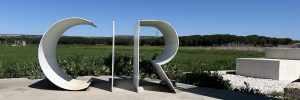 En la imagen se ven las iniciales CIR del Centro de Interpretación de la Resina. El fondo es un campo de trigo que verdea y el cielo es azul.